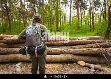 Un homme avec un sac à dos se tient dans la forêt verte devant des bûches coupées. Banque D'Images