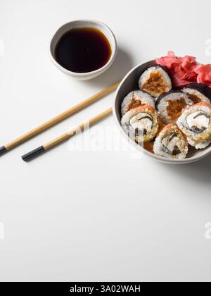 Rouleaux de sushi dans un bol avec du gingembre rose et de la sauce soja et des bâtonnets en bois sur la vue de dessus de table blanche Banque D'Images