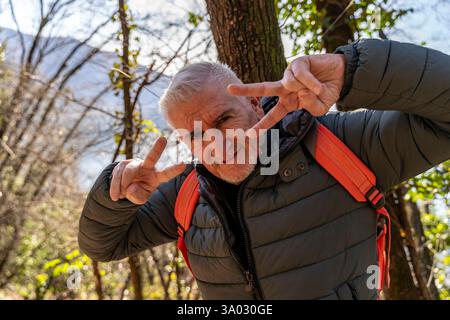 heureux homme d'âge moyen montrant signe de paix avec les mains entourées par la nature en face d'une belle vue de dessus du lac Banque D'Images