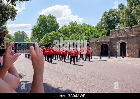 Une bande de gardes britanniques en uniforme rouge et chapeaux en peau d'ours noir défilent dans une rue tandis que les touristes capturent le moment avec des smartphones, Saint Banque D'Images