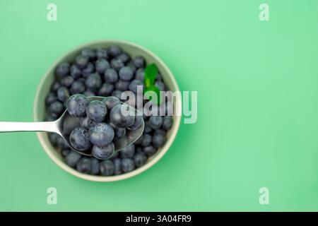 Une cuillerée de bleuets dans un bol. Le bol est blanc et la cuillère est argentée. Les bleuets sont frais et mûrs. Fond vert. Vue de dessus de table Banque D'Images