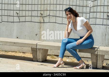 Jeune femme latine profitant d’un moment de paix assise sur un banc en béton contre un mur texturé avec une grille métallique Banque D'Images