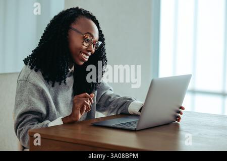Une femme d'affaires afro-américaine souriant tout en travaillant sur son ordinateur portable à un bureau en bois. L'image dépeint un sens du professionnalisme, de la confiance, un Banque D'Images