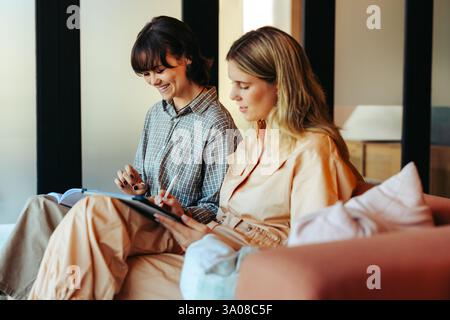 Deux amis étudient ensemble à l'aide d'une tablette et d'un livre, profitant d'une séance productive dans une atmosphère confortable et contemporaine de salon. Banque D'Images