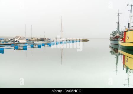 Scène sereine de bateaux amarrés dans un port, capturés par une journée brumeuse avec des reflets d'eau tranquilles, évoquant un sentiment de calme et d'immobilité. Banque D'Images