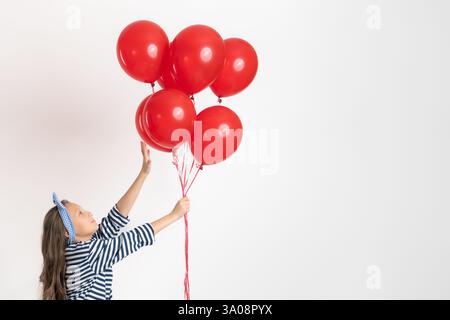 Jeune fille joyeuse tenant dans une main un grand bouquet de ballons rouges, l'autre main atteint des ballons et debout sur la pointe des pieds sur fond blanc. Modèle 10 ans Banque D'Images