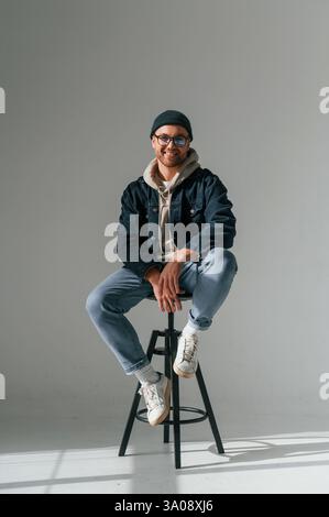 Le mannequin en chapeau et lunettes est assis sur une chaise. Bel homme est dans le studio sur fond blanc. Banque D'Images