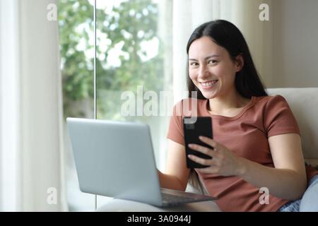 Heureuse femme asiatique posant avec ordinateur portable et téléphone assis sur un canapé à la maison Banque D'Images