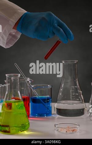 Main gantée tenant un tube à essai contenant du liquide rouge sur divers verreries et produits chimiques de laboratoire, menant des recherches scientifiques. Banque D'Images