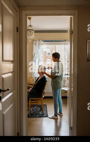 Vue de côté d'une infirmière peignant les cheveux d'une femme âgée à domicile vu à travers l'entrée de la porte Banque D'Images