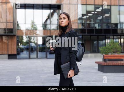 Vue latérale de la jeune femme entrepreneure portant des vêtements noirs avec col haut. Femme d'affaires avec tablette numérique et tasse de café pour aller contre le verre Banque D'Images