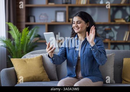 Une jeune femme fait signe pendant un appel vidéo en utilisant une tablette, confortablement assise sur un canapé dans son salon à la maison. Banque D'Images