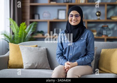 Une jeune femme portant un hijab et des lunettes est assise sur un canapé dans son salon. Elle sourit et regarde la caméra. Banque D'Images