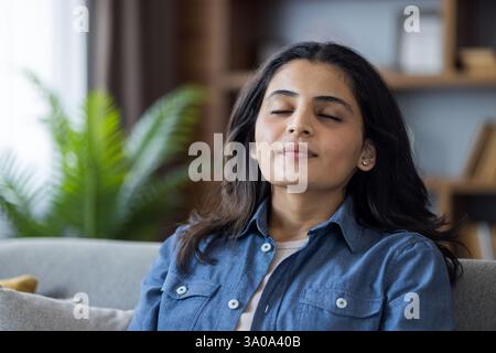 Jeune femme se relaxant à la maison sur le canapé, les yeux fermés, pratiquant la méditation et la pleine conscience pour soulager le stress dans un salon confortable. Banque D'Images