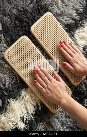 Gros plan des mains avec du vernis à ongles rouge placé sur des planches de massage d'acupressure en bois, sur un tapis texturé gris et blanc. Un concept de bien-être et de détente. Banque D'Images