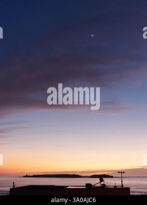 Lune dans le ciel sombre au coucher du soleil avec ciel rouge et île ci-dessous sur la mer Atlantique avec toit en premier plan à Essaouira. 2 mars 2025 Banque D'Images