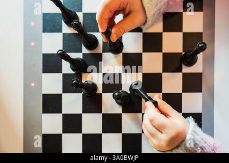 Vue de dessus gros plan des mains de l'enfant jouant aux échecs stratégiques sur un tableau noir et blanc classique, concept d'apprentissage et de divertissement. Banque D'Images