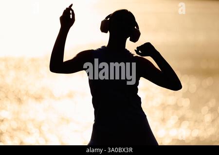 Une jeune belle femme danse librement près de la plage, portant des écouteurs lorsque le soleil se couche. Banque D'Images