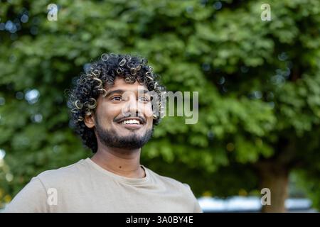 Un jeune homme aux cheveux bouclés et à la barbe courte sourit et lève les yeux sur le fond bokeh d'arbres verts luxuriants. Banque D'Images