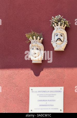 Taormina, Sicile, Italie - 5 juillet 2024 : centre historique. Deux pots de fleurs en céramique blanche, sculptés comme le visage de la femme et ou de l'homme pend contre l'outsi rouge Banque D'Images