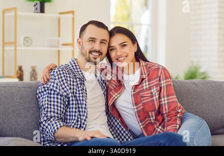 Jeune couple marié heureux assis sur le canapé dans le salon à la maison et souriant. Banque D'Images