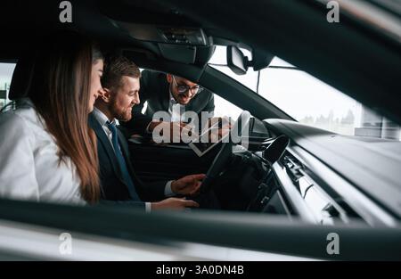 Vue latérale. Le travailleur de salle d'exposition de voiture consulte l'homme et la femme qui sont assis à l'intérieur de l'automobile. Banque D'Images