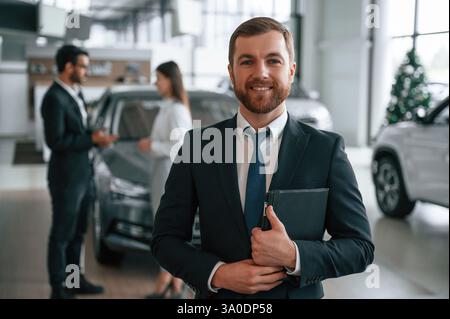 Bel homme est debout et souriant. Trois personnes travaillent ensemble dans le showroom automobile. Banque D'Images