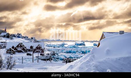 Un paysage hivernal pittoresque avec des maisons traditionnelles entourées de neige, avec des icebergs flottant dans une mer gelée sous un ciel nuageux spectaculaire. Banque D'Images