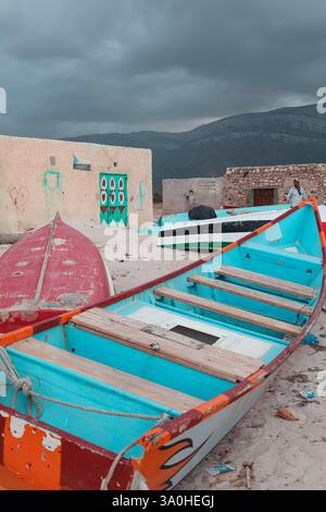 Des bateaux de pêche colorés reposent sur la plage de sable de Socotra, entourés de structures altérées et d'un ciel nuageux spectaculaire. Banque D'Images