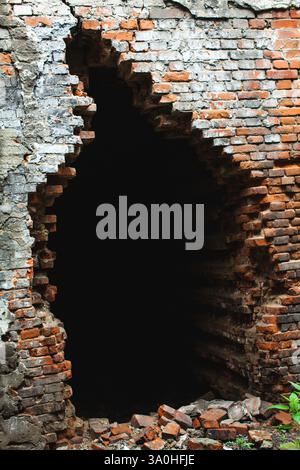 Cette image montre un vieux mur de briques partiellement détruit avec un grand trou au centre, menant à l'obscurité. Les briques rugueuses et en ruine, varyi Banque D'Images