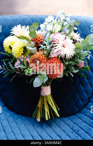 Bouquet de mariage avec dahlias orange, gerberas roses et blanches, fleurs jaunes et bleu clair couchées sur un fauteuil matelassé bleu Banque D'Images
