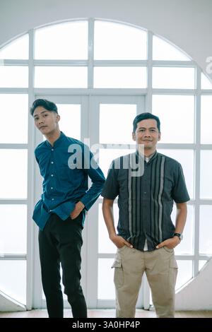 Deux hommes adultes joyeux, vêtus d'une tenue décontractée moderne, souriant devant une fenêtre lumineuse. L'image montre une relation amicale et confiante wi Banque D'Images