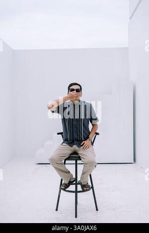 Un jeune homme aux lunettes sombres assis dans un studio minimaliste moderne, la main à la bouche dans un geste de silence. Le fond blanc propre et le mépris Banque D'Images