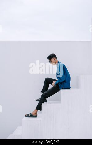 Un jeune homme élégant, vêtu d'une chemise bleue élégante et d'un pantalon sombre, s'assoit soigneusement sur des marches blanches. L'image est un mélange de la mode moderne a Banque D'Images