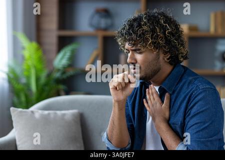 Homme aux cheveux bouclés toussant assis sur un canapé dans un cadre intérieur, présentant éventuellement des symptômes de maladie ou d'allergie. Banque D'Images
