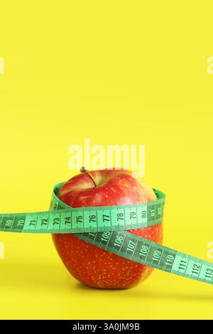 Ruban à mesurer enroulé autour d'une pomme. Métaphore - figure ronde. Pomme rouge mûre et ruban à mesurer sur fond jaune. Concept de saine alimentation, régime alimentaire Banque D'Images