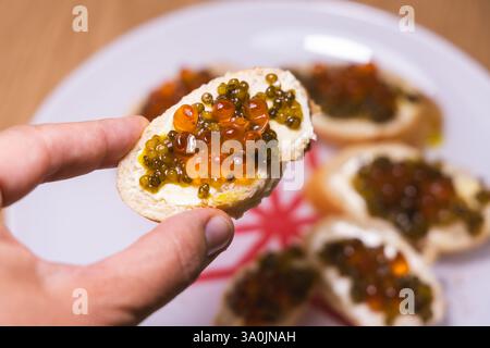 Un gros plan d'une main tenant un morceau de bruschetta surmonté de caviar rouge et vert sur tartinade crémeuse, avec plus d'apéritifs sur une assiette dans le backgroun Banque D'Images