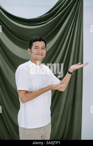 Un homme souriant dans un t-shirt blanc présente ou introduit quelque chose avec un geste invitant de la main. Il semble confiant et amical contre un ol Banque D'Images
