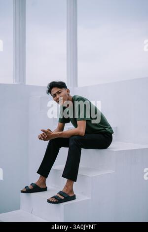 Un jeune homme élégant est assis sur des escaliers blancs près de colonnes architecturales dans une pose réfléchie. Le cadre évoque un sentiment d'élégance minimaliste et moderne Banque D'Images