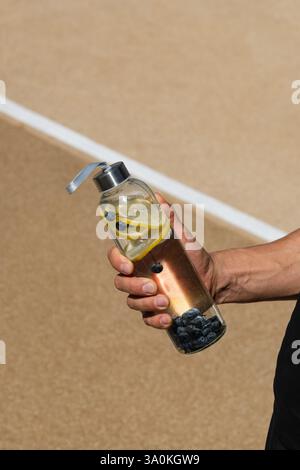 Bouteille réutilisable en verre sport avec citron et baie ou isotonique dans la main de l'athlète. Concept détox, eau potable. Suivi quotidien de l'eau. Bilan eau pour WO Banque D'Images