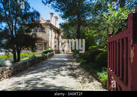 Un sentier calme et ensoleillé menant à une charmante villa, encadrée par une végétation luxuriante et de grands arbres, avec un portail rouge rustique invitant un retre paisible et privé Banque D'Images