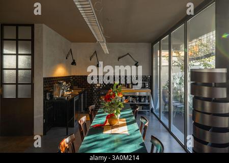 Une cuisine élégante avec un dosseret aux tons foncés, un éclairage minimaliste et une table à manger rustique décorée de fleurs de fête. Les grandes fenêtres en verre apportent la lumière naturelle, créant une atmosphère chaleureuse et accueillante avec un mélange d'esthétique moderne et vintage Banque D'Images