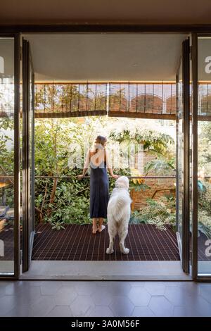 Une femme dans une robe minimaliste se tient avec son grand chien blanc sur un balcon moderne, donnant sur une végétation luxuriante. Les portes vitrées ouvertes allient confort intérieur et sérénité extérieure, évoquant la paix et la pleine conscience Banque D'Images