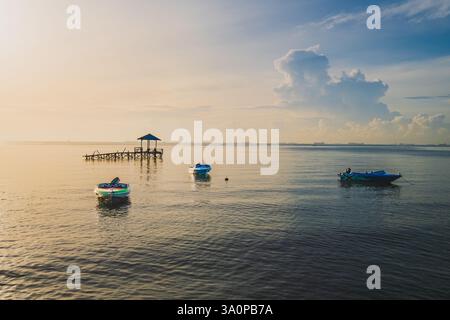 Un paysage marin serein avec trois petits bateaux ancrés dans une eau calme. Une jetée en bois avec un gazebo s'étend dans la mer sous un ciel doux et nuageux. Banque D'Images