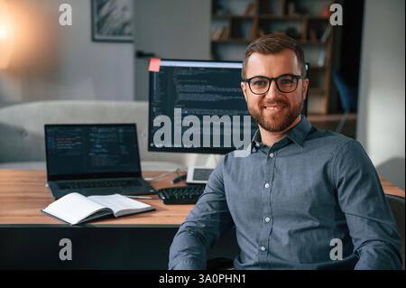 Le développeur de logiciels est assis et pose. Homme en vêtements formels travaille dans le bureau moderne. À l'aide de l'ordinateur. Banque D'Images