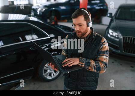 Debout avec l'ordinateur portable dans les mains et dans les écouteurs. L'homme en uniforme travaille dans le salon automobile. Banque D'Images