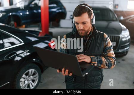 Debout avec l'ordinateur portable dans les mains et dans les écouteurs. L'homme en uniforme travaille dans le salon automobile. Banque D'Images