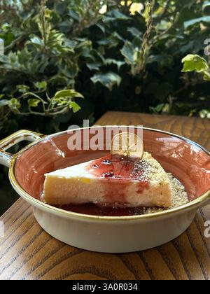 Une tranche de cheesecake crémeux garnie de sauce aux baies rouges et d’une garniture de citron séché, servie dans un plat en céramique sur une table en bois avec un fond vert Banque D'Images