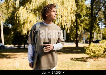Jeune homme se promène dans un campus ensoleillé tenant un café et regardant soigneusement devant l'avant. Banque D'Images