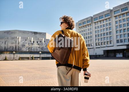 Un jeune homme à la mode se promène dans une zone urbaine, profitant de son café et du soleil éclatant. Banque D'Images